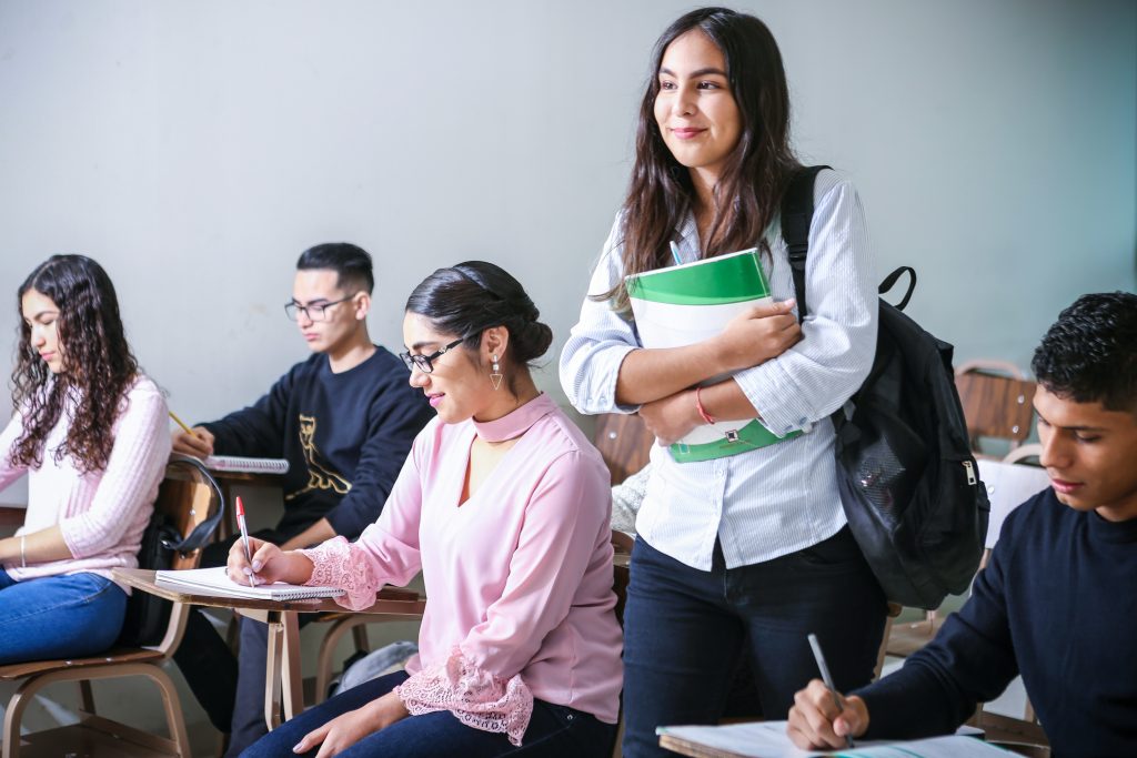 Smiling female student carrying a green notebook and backpack while walking into a classroom with other students taking notes.